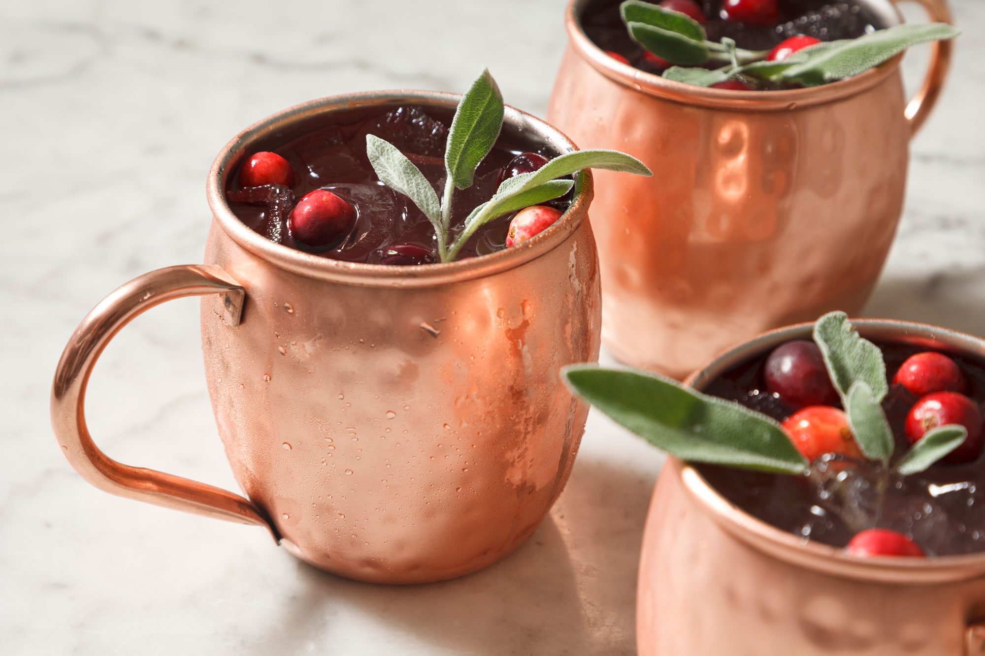 3/4th shot of cranberry moscow mule garnished with frozen cranberries and fresh sage in a copper mug