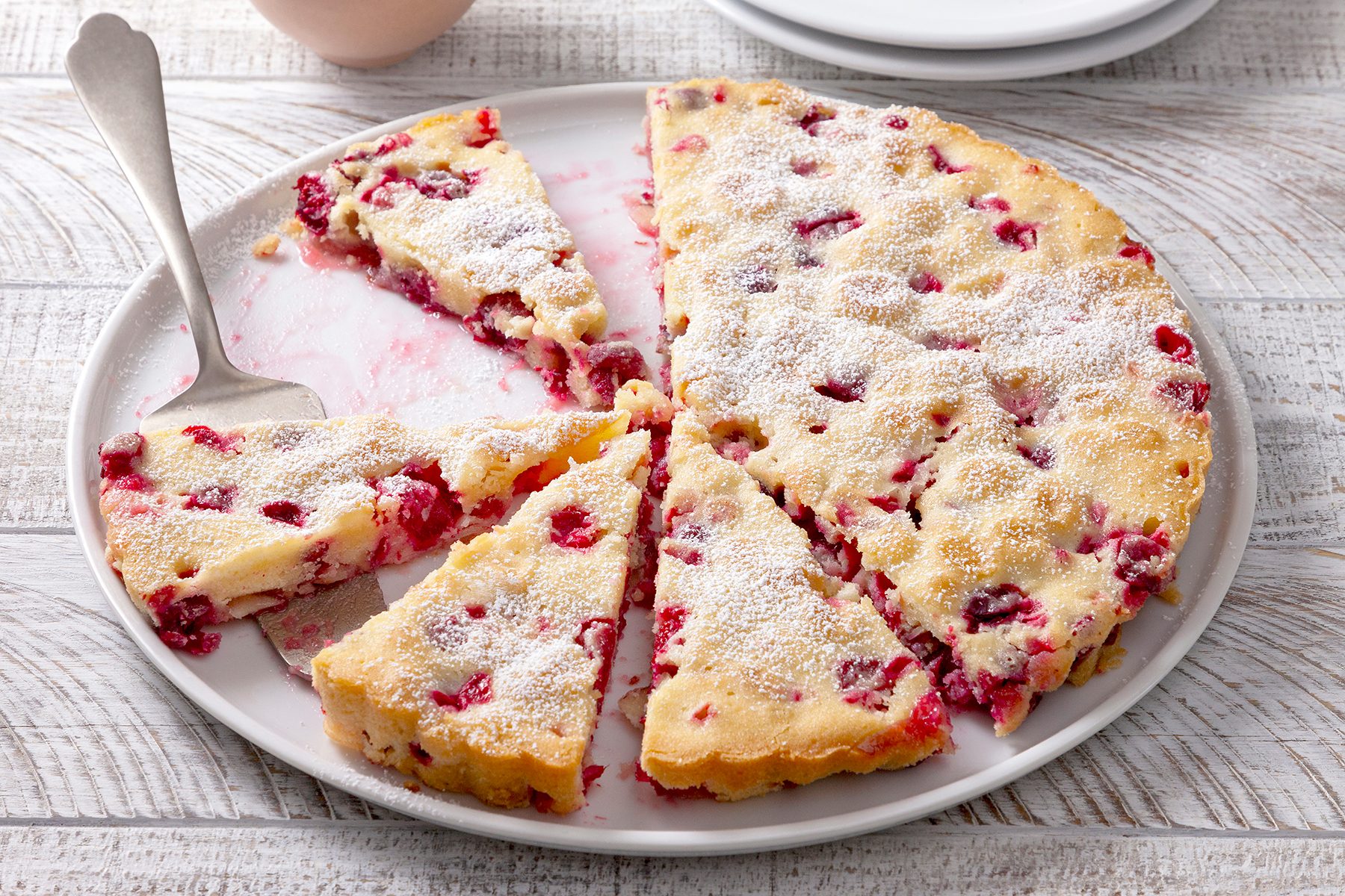 A sliced berry cake with powdered sugar on top is served on a white plate. A silver serving spatula rests on the plate. The background is a light wooden table.
