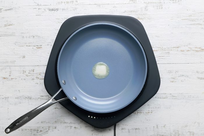overhead shot; white wooden background; a blue frying pan with a silver handle, placed on a black square plate, Inside the pan, there’s a small pat of butter melting in the center