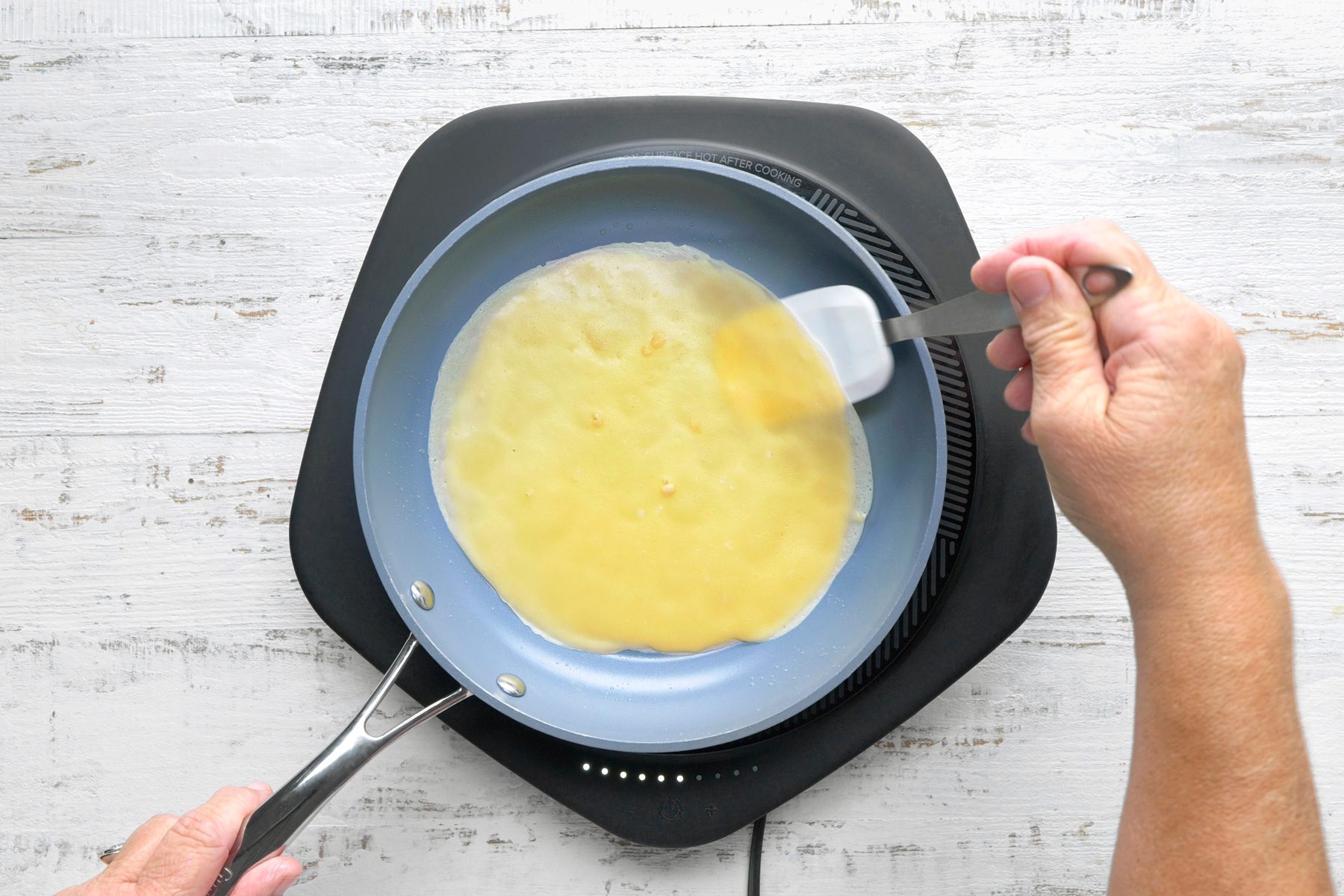 a top-down view of a person cooking a pancake in a blue non-stick skillet, The batter is spreading evenly across the bottom of the skillet, and the edges are starting to set, The person is holding a spatula in one hand and is using it to smooth out the batter and prevent it from sticking to the skillet. The skillet is placed on a black electric burner
