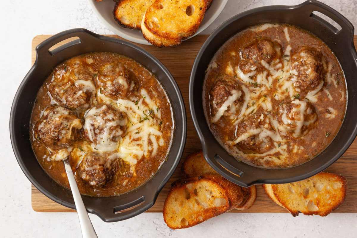 Two bowls of Taste of Home French Onion Meatballs in black bowls on a wooden cutting board, with toasted baguette slices