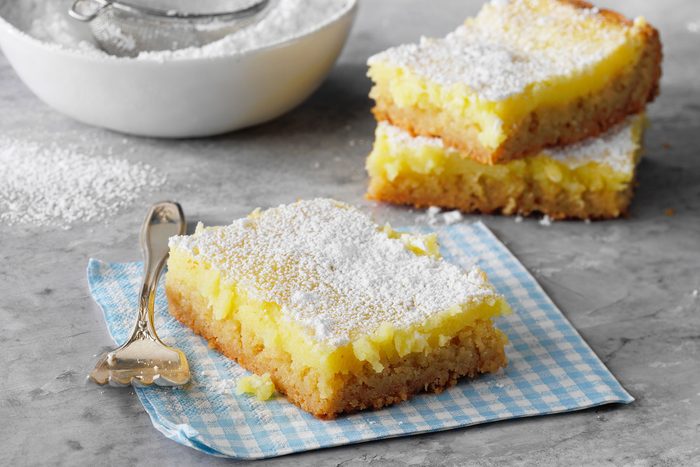 Two lemon bars topped with powdered sugar rest on a blue gingham napkin. A fork is placed beside them. In the background, a bowl of powdered sugar with a sifter sits on a gray surface.