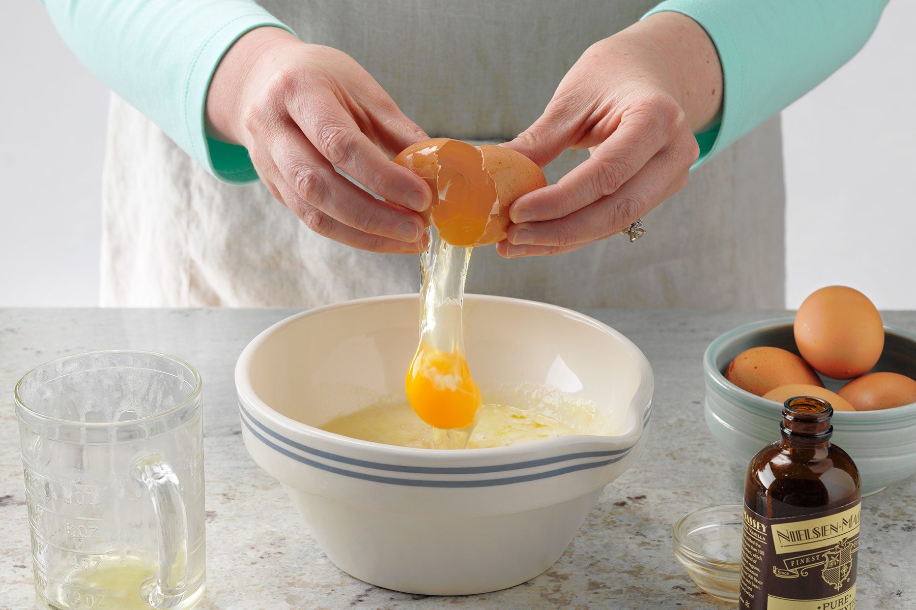 A person cracking an egg into a mixing bowl on a kitchen counter. Surrounding the bowl are a glass measuring cup, a bottle of vanilla extract, and a small bowl filled with eggs. The person is wearing a light blue sleeve.