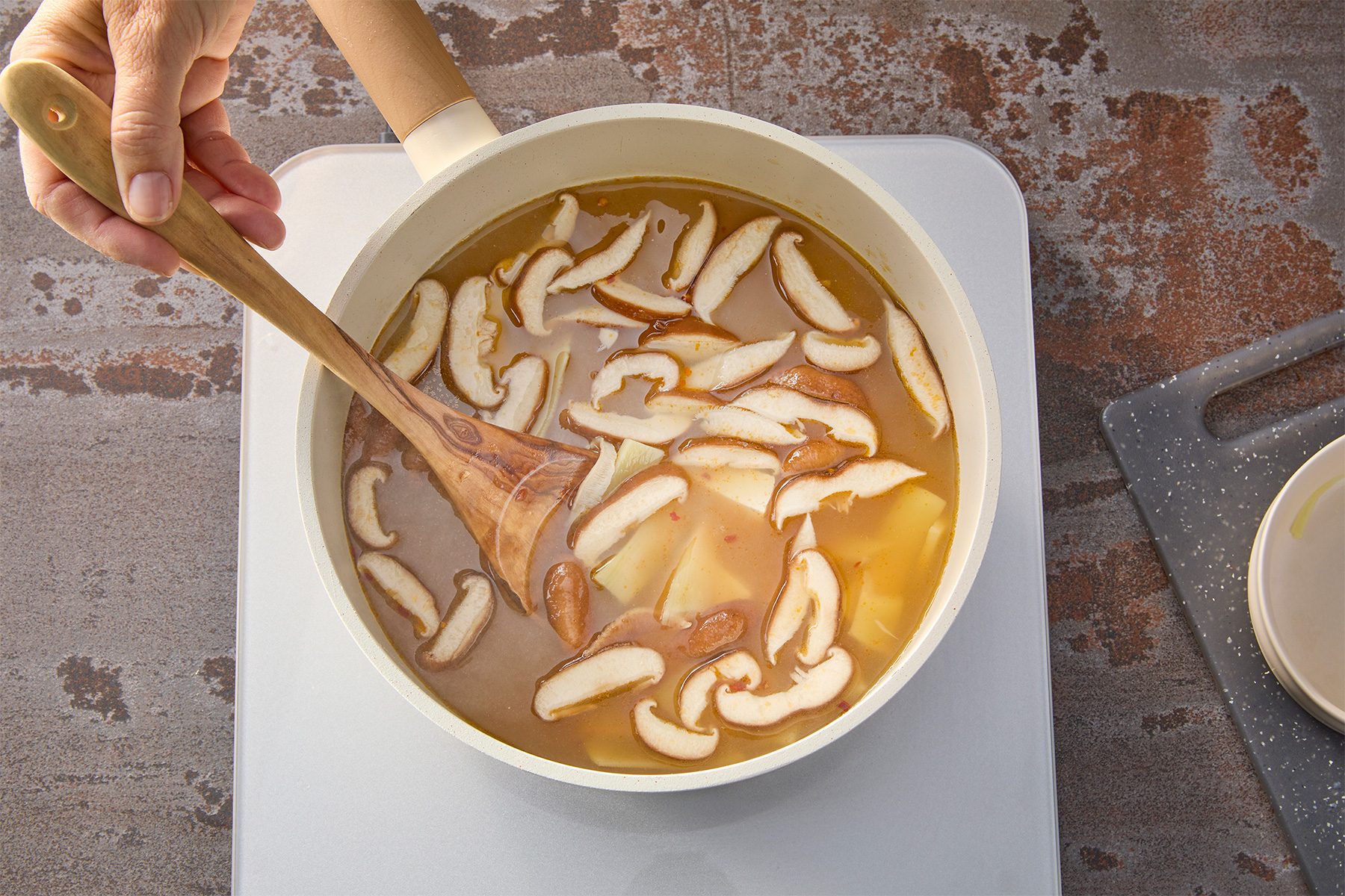 Overhead shot of add tofu; mushrooms and bamboo shoots; cook 10-15 minutes or until mushrooms are soft; wooden spoon; brown texture background;