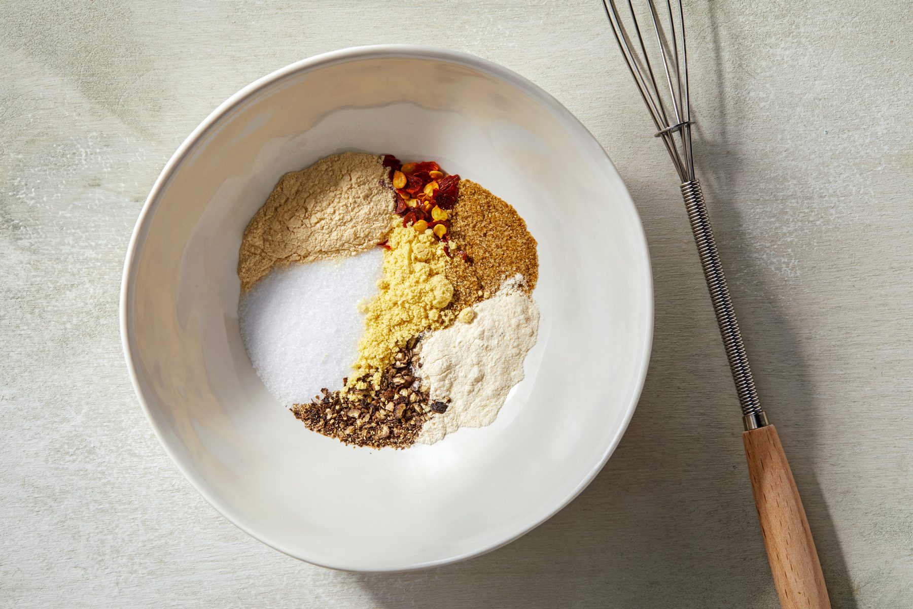 Overhead shot of a large bowl filled with a mixture of salt, garlic powder, pepper, mustard, onion powder, celery salt, and red pepper flakes, with a whisk positioned beside the bowl.