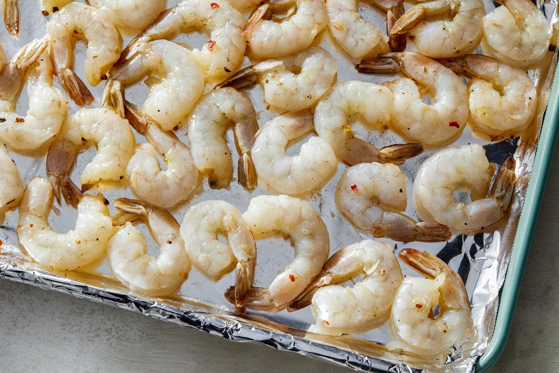 3/4 shot of a baking sheet lined with aluminum foil, featuring a large number of raw shrimp arranged and spaced apart, coated in a mixture of spices.
