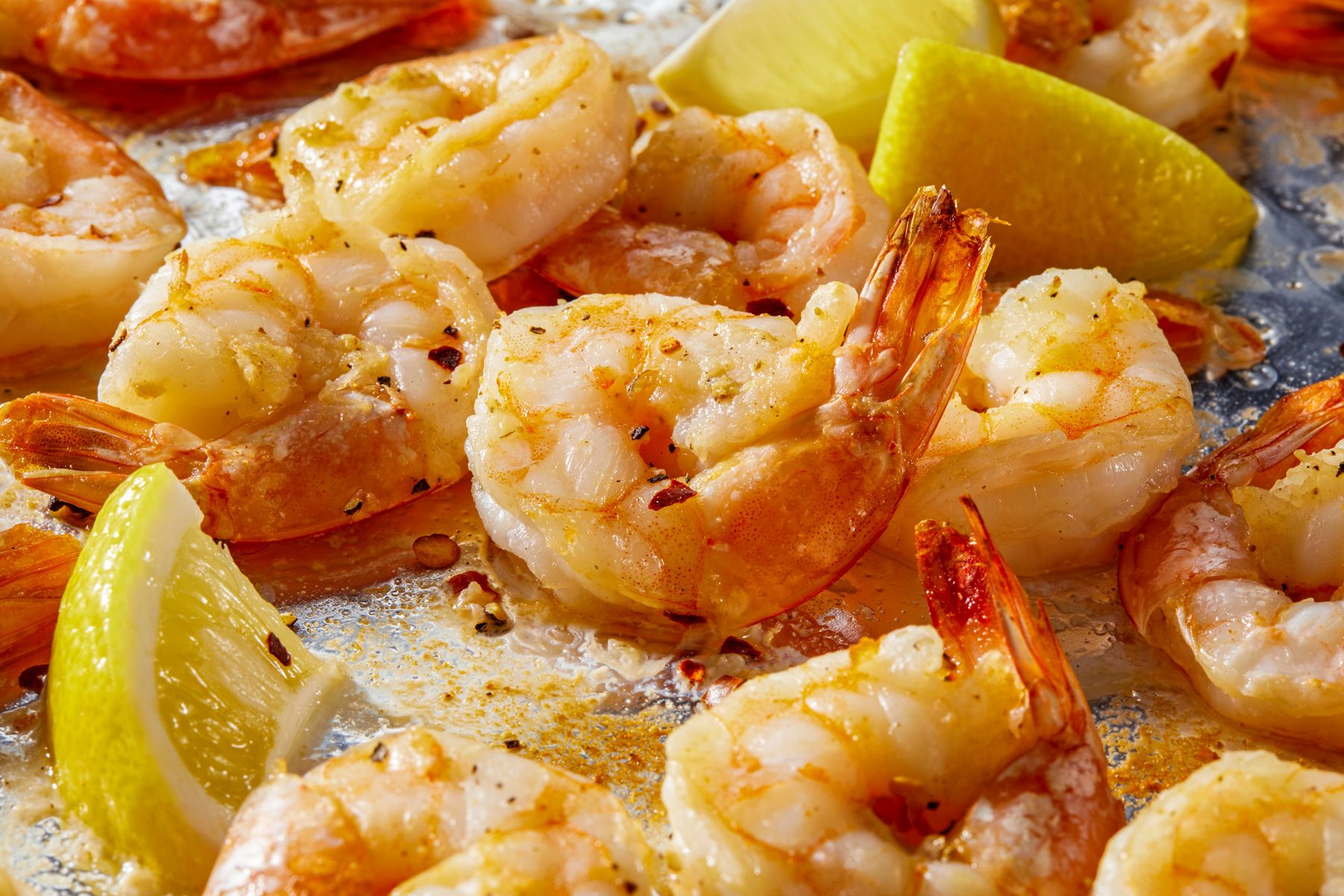 Horizontal push shot of a baking sheet filled with cooked shrimp in a pale pink color, scattered across the sheet, with slices of lemon placed among them.