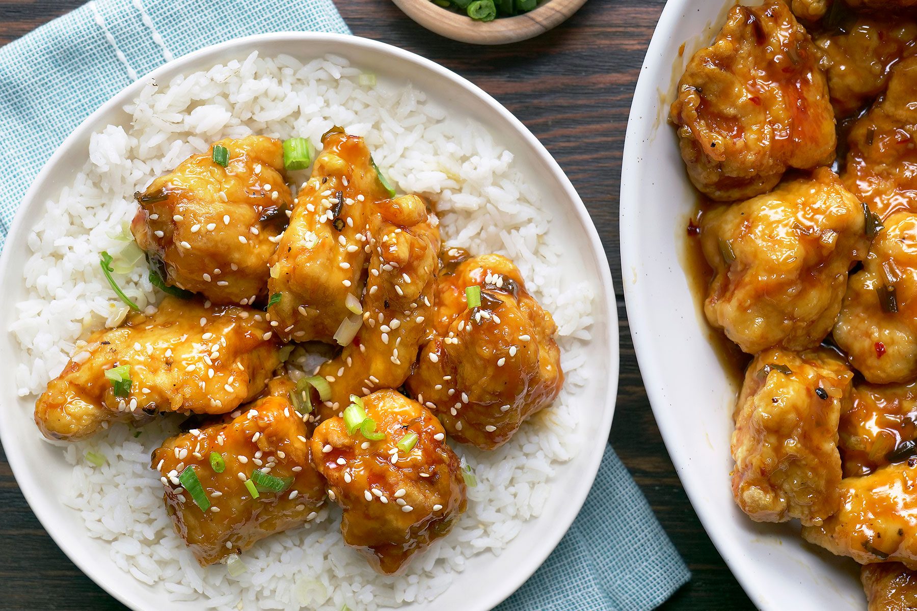A plate of white rice topped with pieces of golden-brown fried chicken glazed in a sticky sauce, garnished with sesame seeds and chopped green onions. A portion of the dish is also visible in a large serving bowl nearby.