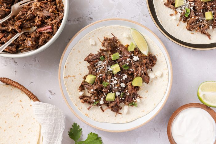 Instant pot beef carnitas served on flour tortillas, garnished with queso fresco, fresh cilantro and cubed avocado.