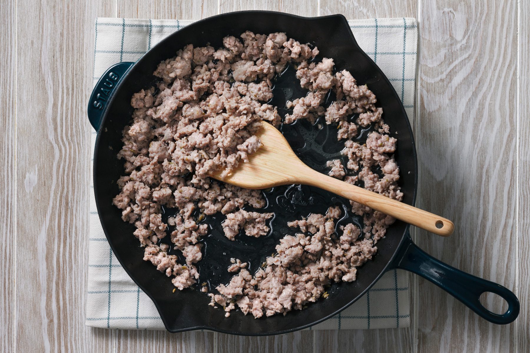 Overhead shot of a cast iron skillet with a blue handle sits on a white checkered dishtowel on a light-colored wooden table ; the skillet is filled with browned ground meat and a wooden spoon lays across the meat.