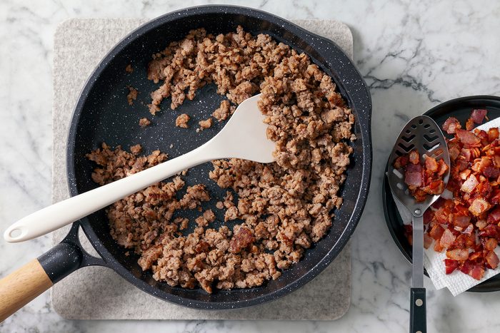 A skillet on a marble surface contains browned ground sausage. A white spatula rests in the skillet. To the right, cooked bacon pieces are in a black bowl with a slotted spoon resting on top.
