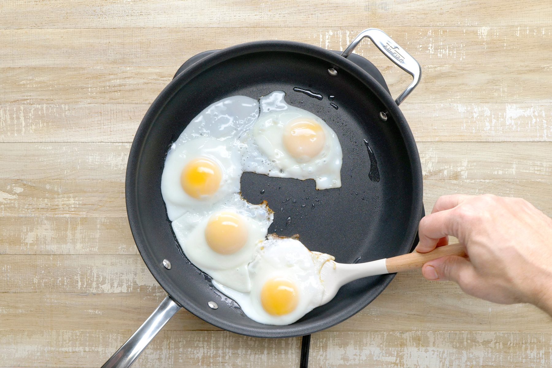 overhead shot of a non-stick skillet filled with oil, Four fried eggs are cooking in the oil, with the yolks still runny, A wooden spatula is being used to gently lift one of the eggs, revealing the cooked white underneath;