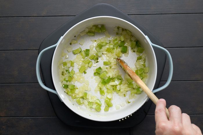A person stirs diced onions and green bell peppers in a white pot on a black stovetop with a wooden spoon. The vegetables are being sautéed, showing a mix of translucent and vibrant colors.