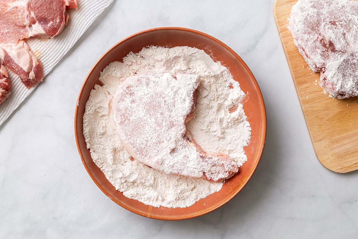 Pork chop being coated in flour in a shallow dish for step one of Maple-Glazed Pork Chops for Taste of Home