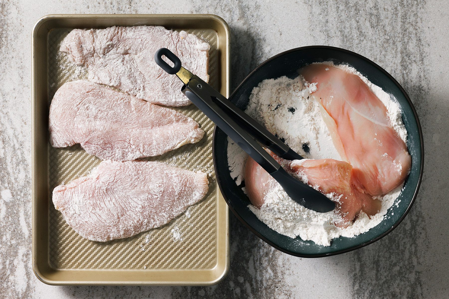 overhead shot of four breaded chicken breasts in a baking dish and a bowl of flour, There are also tongs in the image;