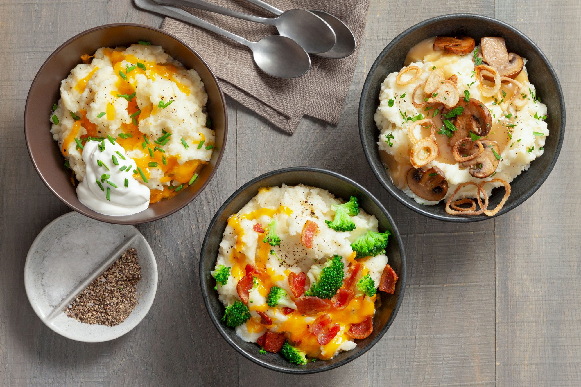 Overhead shot of Mashed Potato Bar; serve in three bowls; spoons; salt and pepper; napkin; wooden surface;