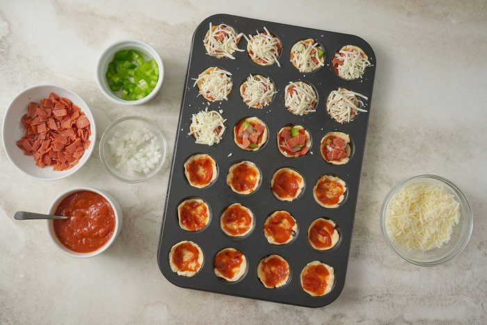 Overhead view of pizza sauce, chopped onion, green pepper, turkey pepperoni, and shredded mozzarella being added to the dough cups for the Taste of Home Pizza Bites recipe.