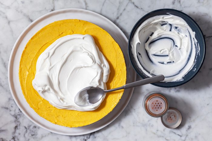 Overhead shot of a pumpkin cheesecake on a plate, The cheesecake is a vibrant orange color and is topped with a layer of whipped cream, A spoon is scooping the whipped cream to create swirls on top of the cheesecake, A bowl of whipped cream is visible in the background, There are also two shakers of spices, likely cinnamon and nutmeg, placed next to the cheesecake.
