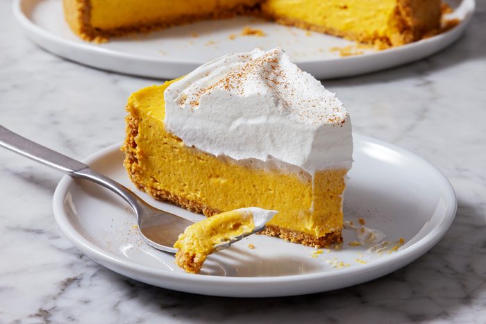 3/4th shot of a slice of pumpkin cheesecake on a white plate, The cheesecake is a vibrant orange color and is topped with a layer of whipped cream, A sprinkle of cinnamon is dusted over the whipped cream, A fork is placed next to the slice of cheesecake.