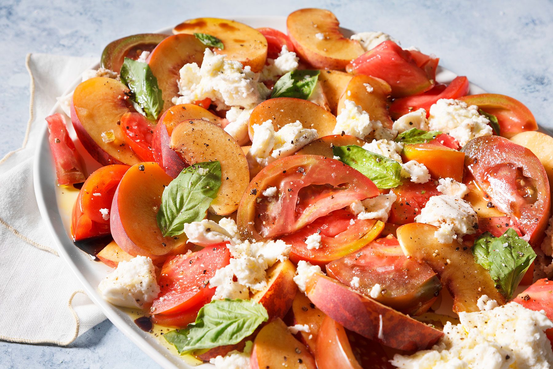A fresh salad featuring sliced tomatoes and peaches, topped with dollops of creamy burrata cheese and scattered basil leaves, seasoned with black pepper, and drizzled with olive oil on a white plate.
