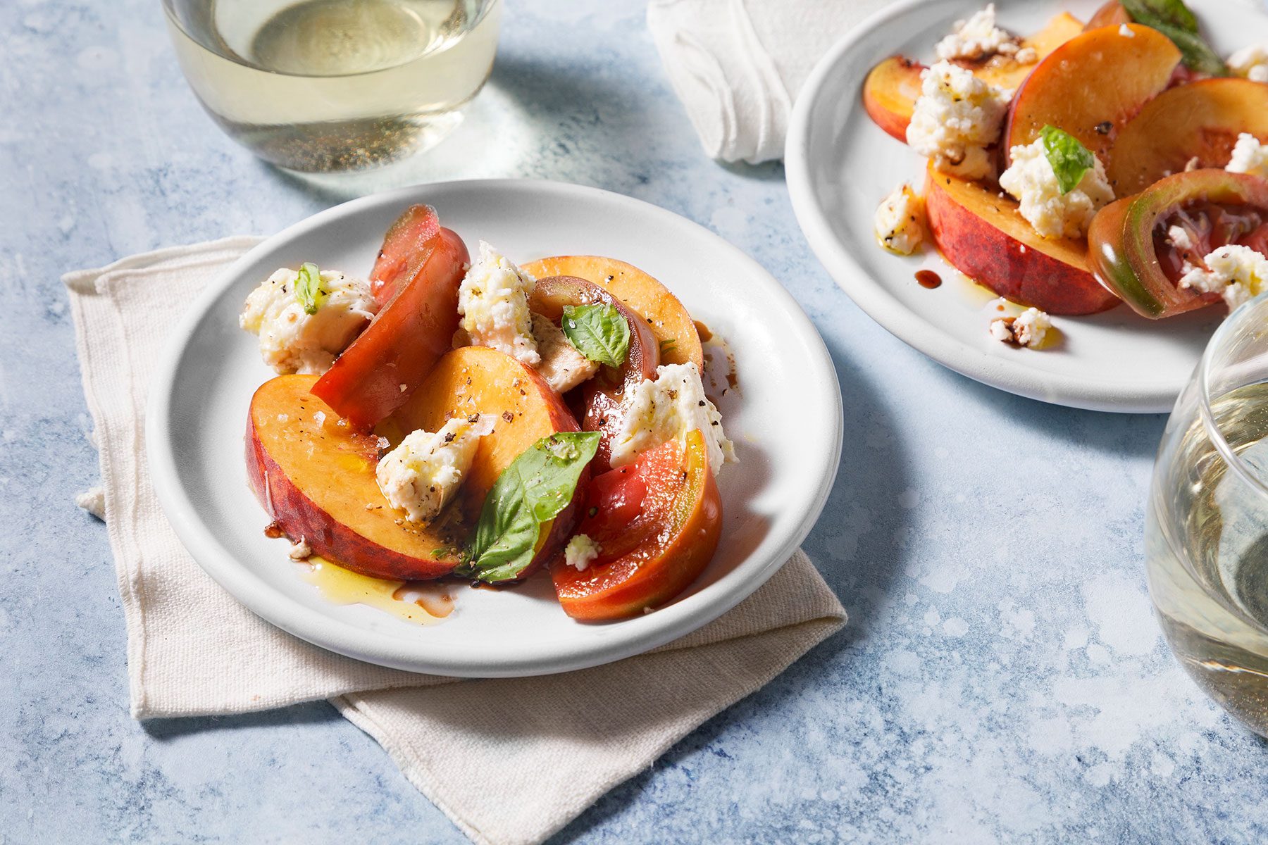 A fresh salad on a white plate features sliced peaches, tomatoes, crumbled cheese, and basil leaves, all drizzled with a balsamic glaze. The dish is set on a cloth napkin, with a glass of white wine in the background.