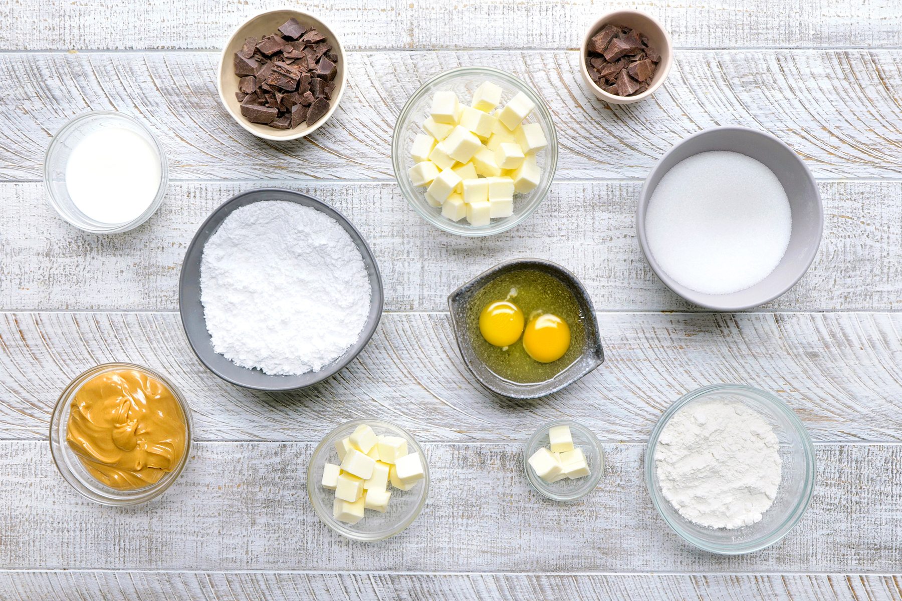 A flat lay of baking ingredients on a wooden surface: small bowls with cubed butter, chocolate pieces, granulated sugar, powdered sugar, eggs, peanut butter, flour, and milk.