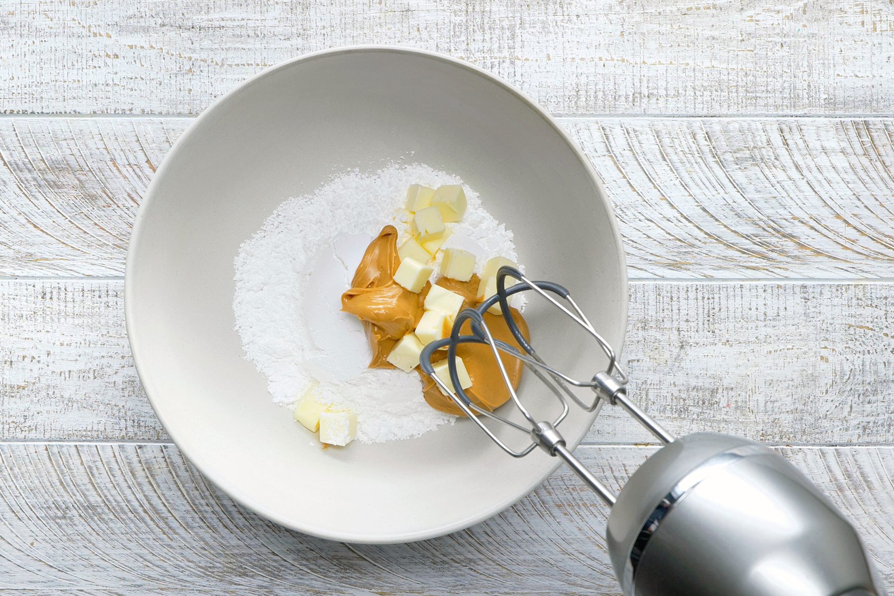 A mixing bowl on a wooden surface containing peanut butter, softened butter, and powdered sugar. A hand mixer is positioned on the right side, ready for mixing the ingredients.