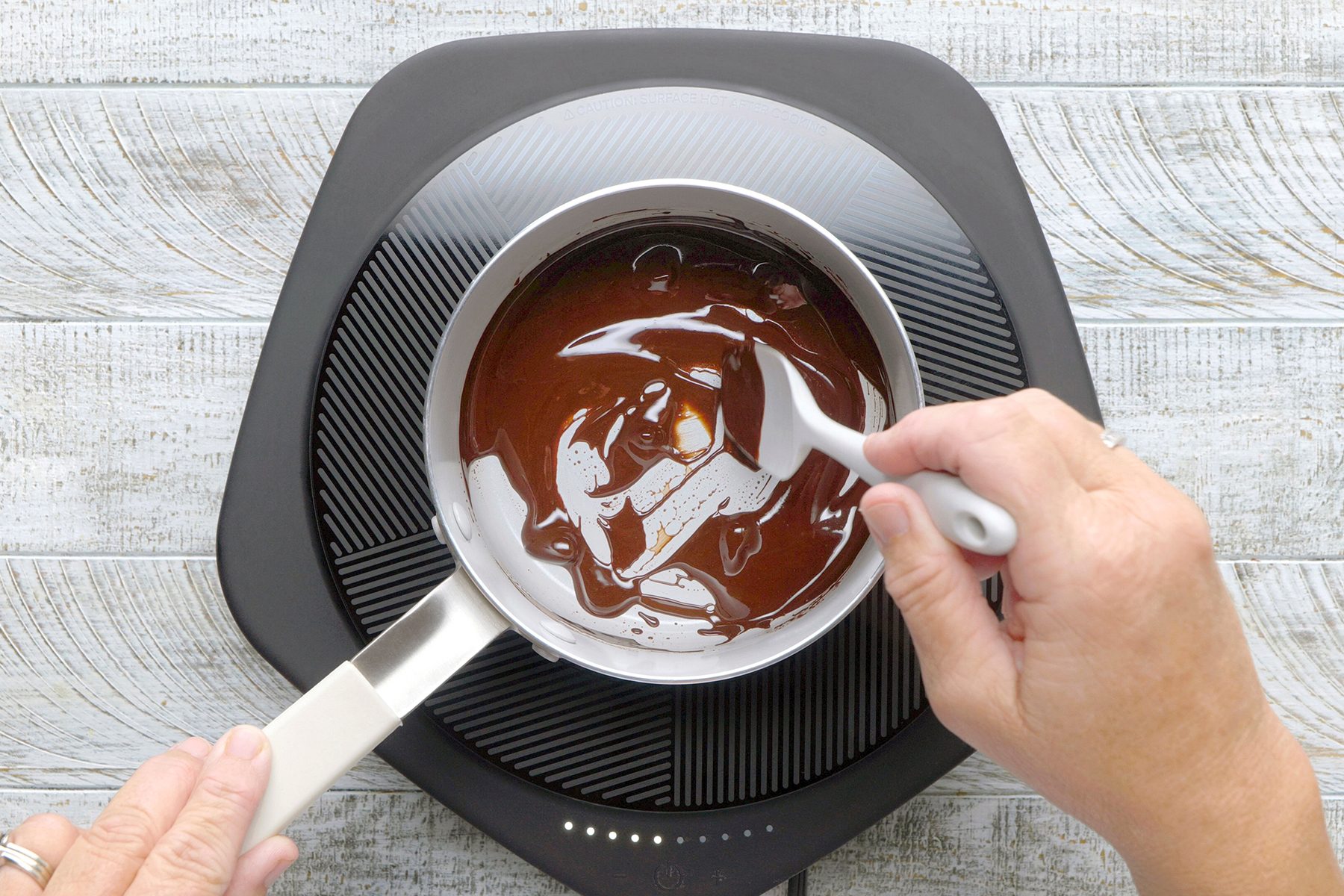 A person stirs melted chocolate in a small saucepan on an induction cooktop. The background is a light, textured wooden surface.