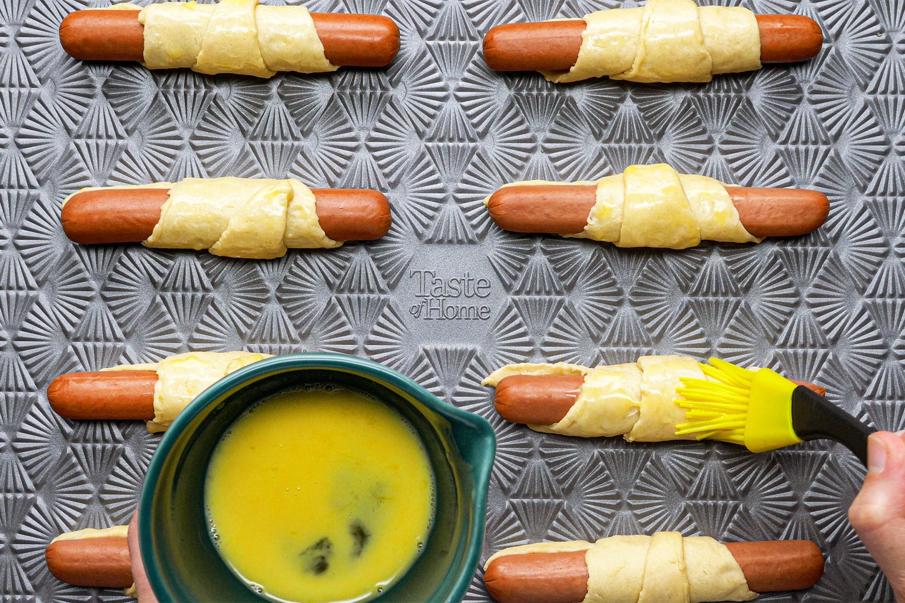 overhead shot; white wooden textured background; brushing egg and water; over rolls; hot dogs; at wide ends; of triangles and rolled up; over baking tray;