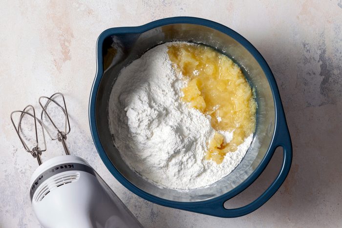 A large mixing bowl contains a mixture of angel food cake mix and crushed pineapple with juice. The ingredients are being beaten together on medium-low speed until fully combined.