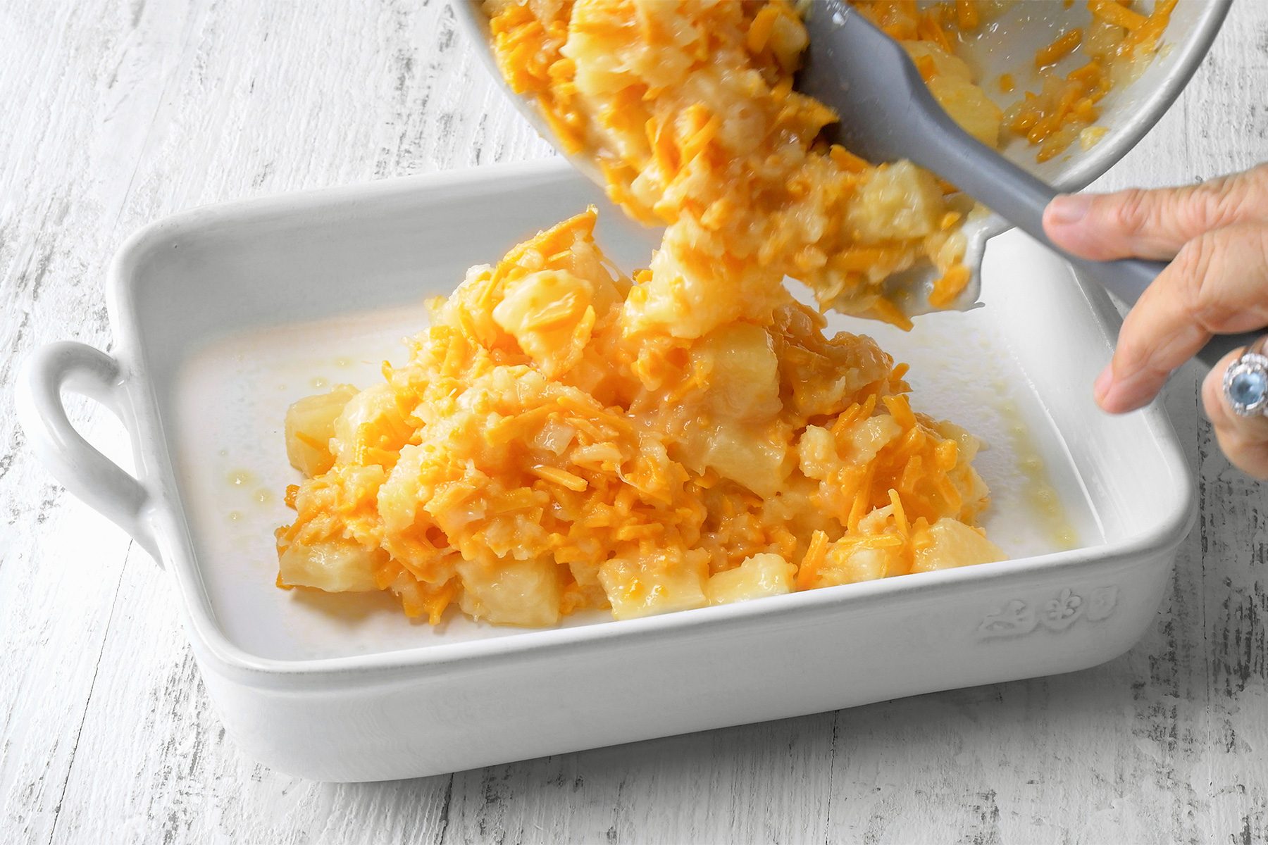 3/4th shot of a white baking dish filled with a creamy, golden-yellow mixture, A person's hand is pouring crushed crackers onto the top of the mixture, The crackers are creating a golden brown topping, The baking dish is sitting on a wooden surface;