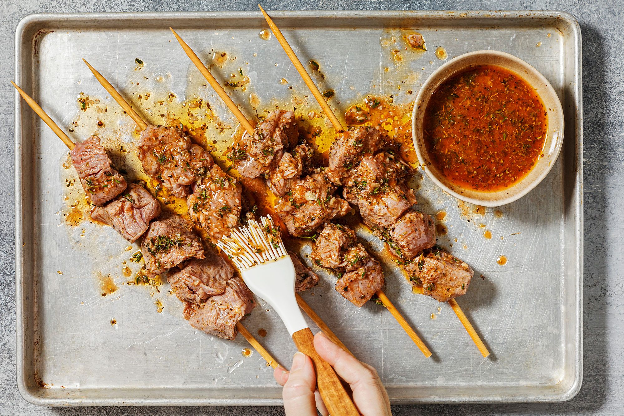 overhead shot of a baking sheet filled with lamb skewers, The skewers are made of metal rods with cubes of lamb meat threaded onto them, The meat is marinated in a reddish-brown sauce, A small bowl of the marinade is visible in the background, and a pastry brush is being used to brush the marinade onto the skewers;