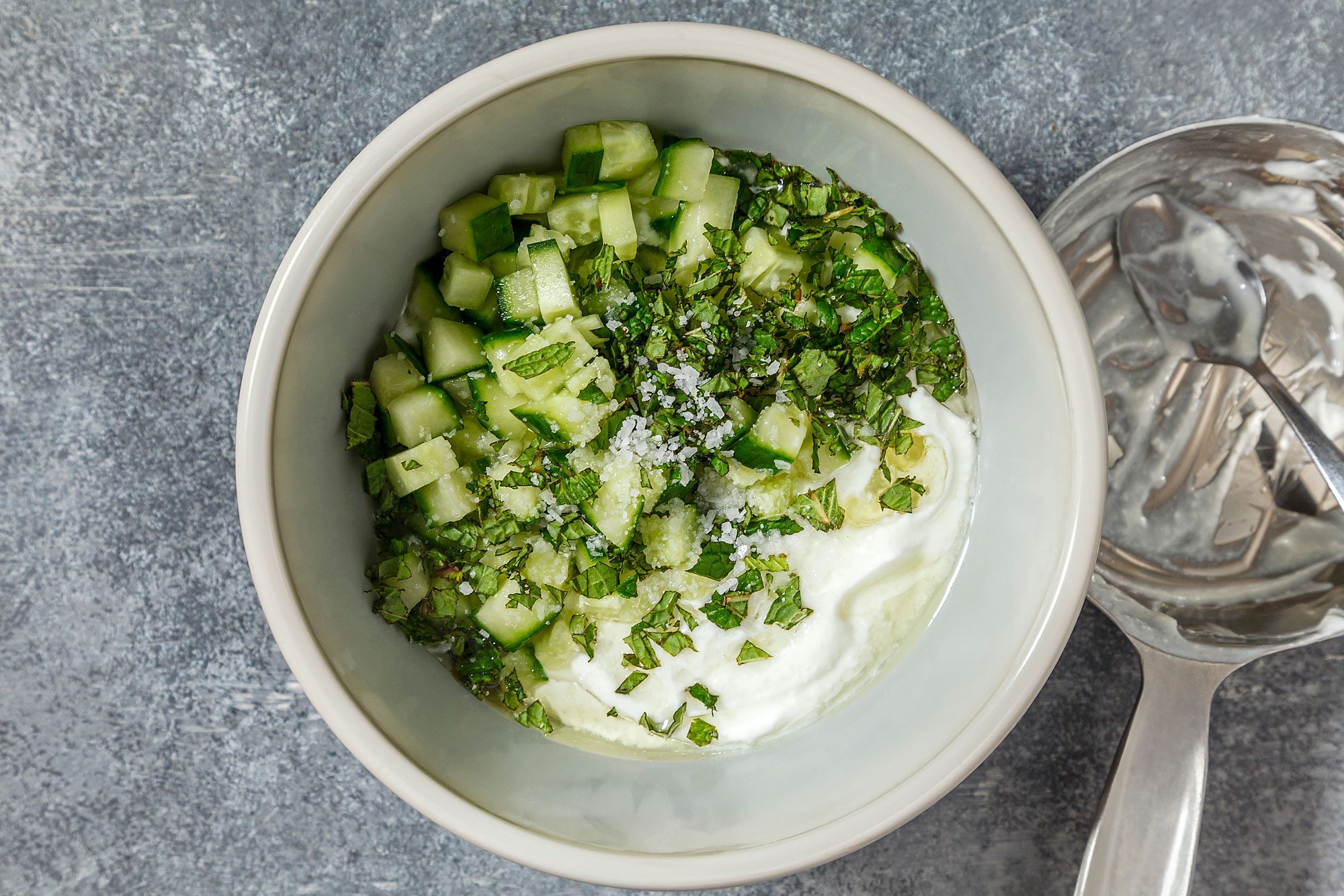 overhead shot of a white bowl contains a refreshing salad of diced cucumbers, chopped mint, and yogurt, The cucumbers are a vibrant green color, the mint leaves are bright green, and the yogurt is creamy white;