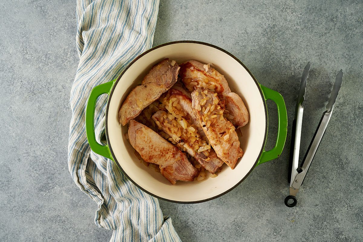 Overhead view of country-style pork ribs and onions browning in a Dutch oven with canola oil for the Taste of Home Pork and Sauerkraut recipe.