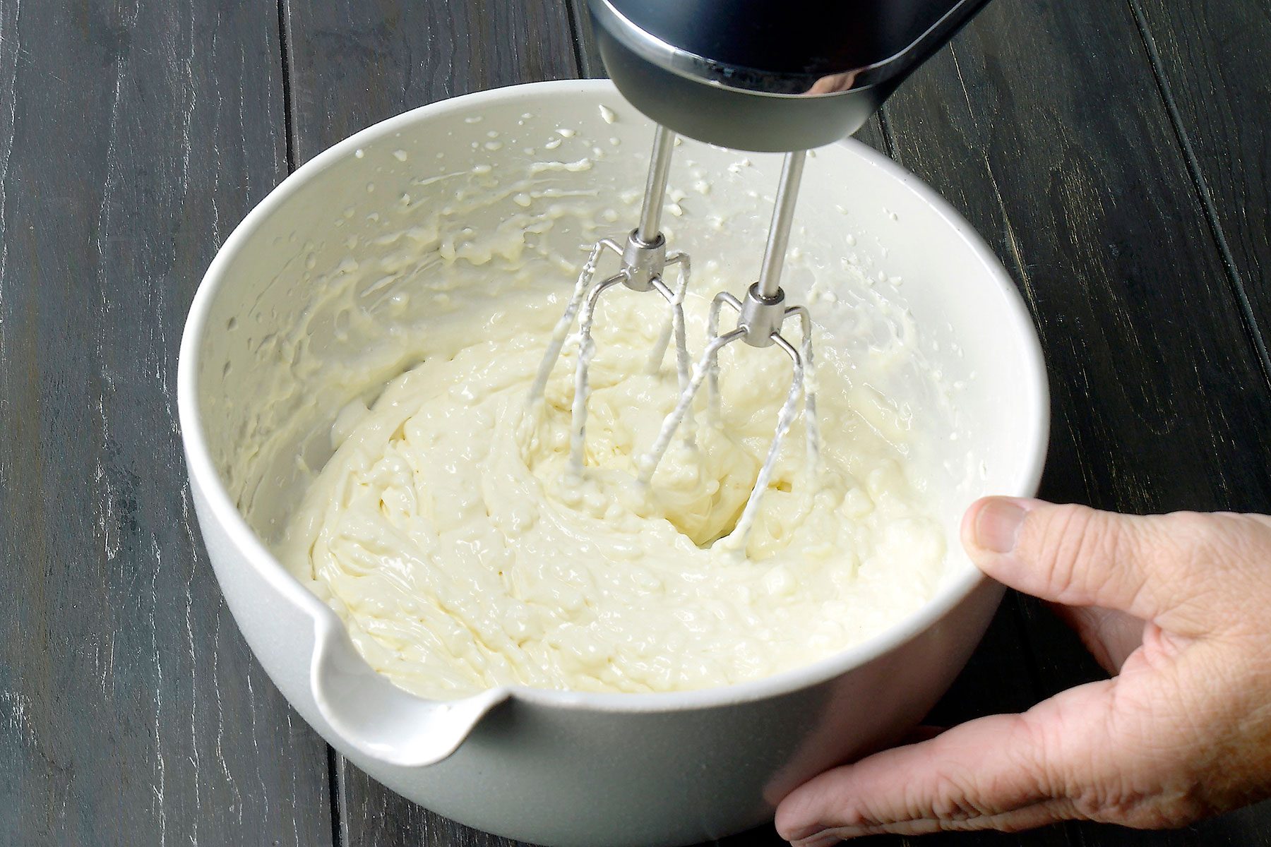 A person's hand holds a white mixing bowl while using a handheld electric mixer to blend a creamy batter. The bowl rests on a dark wooden surface.