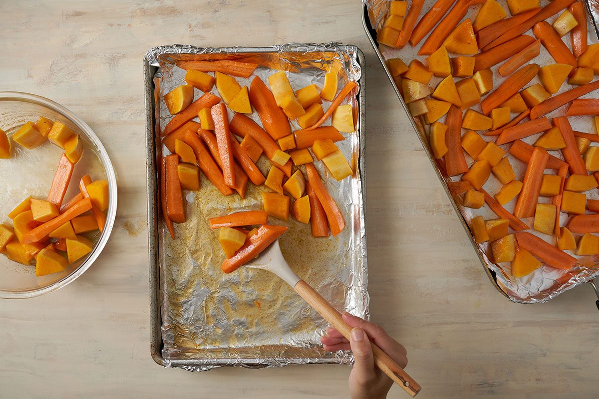 Overhead view of seasoned butternut squash and carrots spread on foil-lined baking pans, roasting in the oven for the Taste of Home Roasted Vegetables with Walnuts recipe.