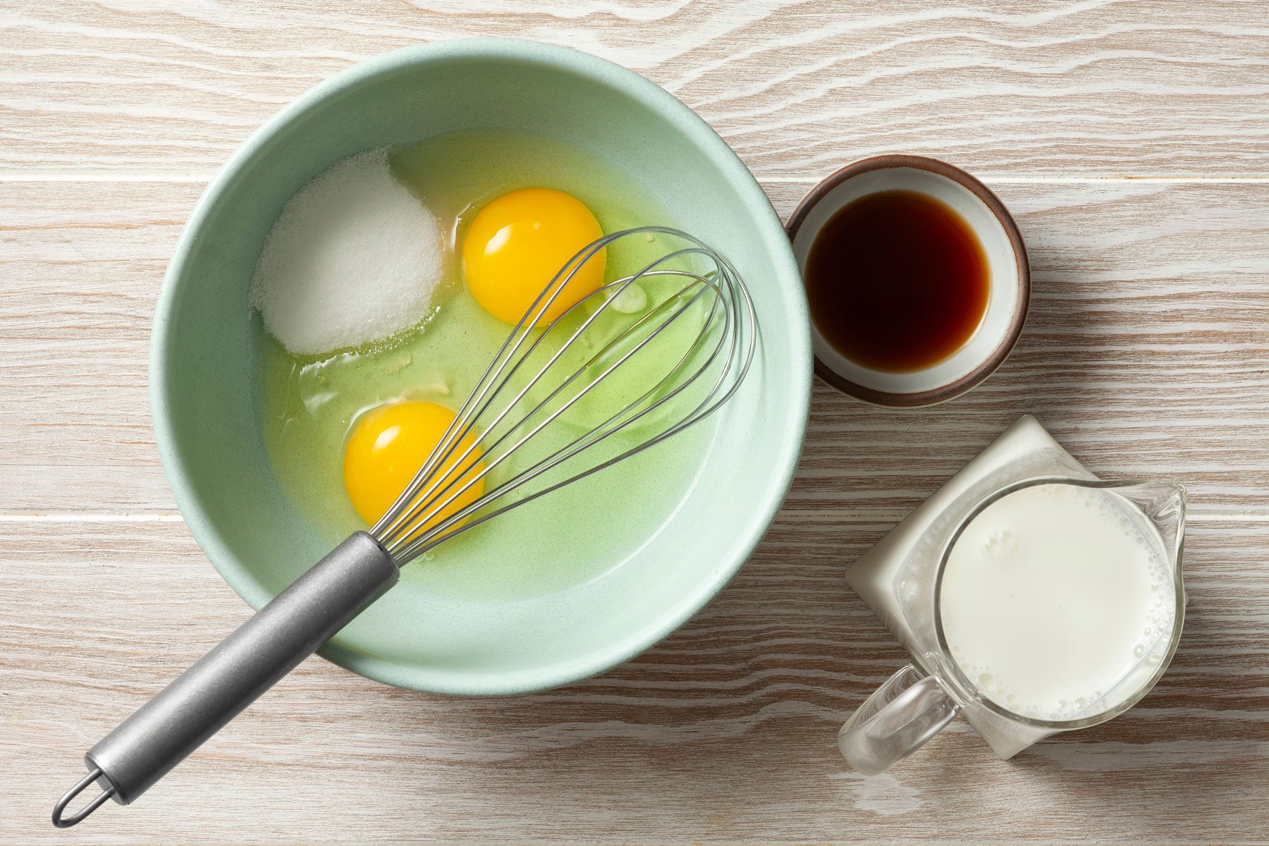Eggs and granulated sugar in a small bowl with vanilla and milk next to it.