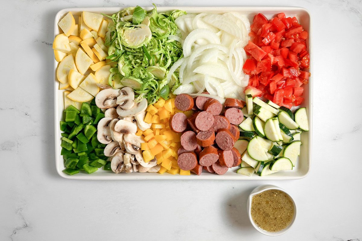 Ingredients for Taste of Home Sheet Pan Sausage and Veggies on a white baking sheet next to a cup of vinaigrette