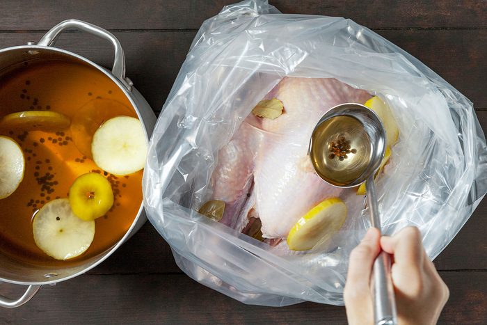A raw turkey in a clear plastic bag being brined with a liquid containing apple slices and spices. A hand pours the brine from a pot using a ladle. The setup is on a dark wooden surface.