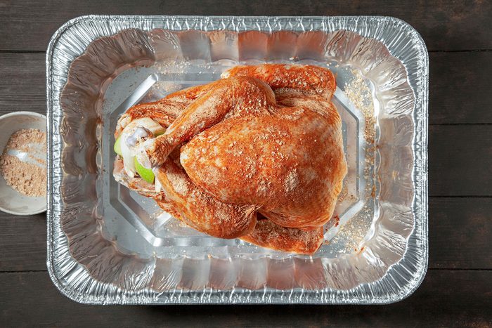 A raw, seasoned whole turkey placed in a large, rectangular aluminum roasting pan, ready for cooking. The turkey is sprinkled with spices, and a small bowl of seasoning is visible beside the pan on a dark wooden surface.