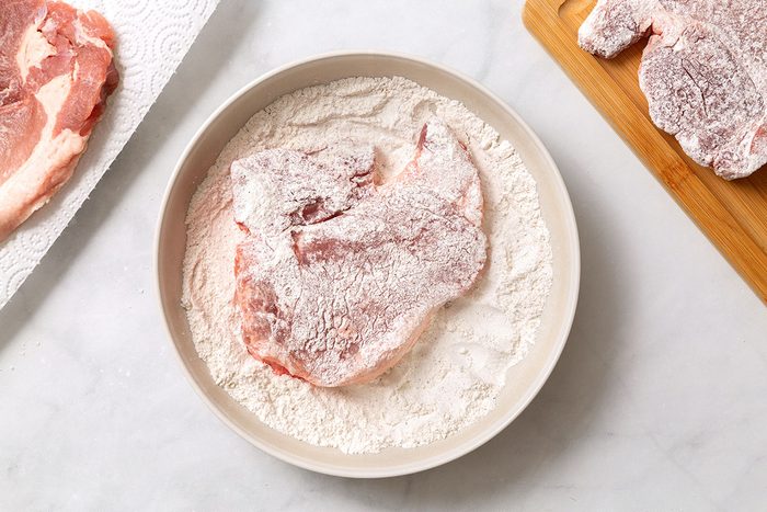 Pork chop coated in flour mixture in a shallow bowl for step one of Smothered Pork Chops in Oven for Taste of Home