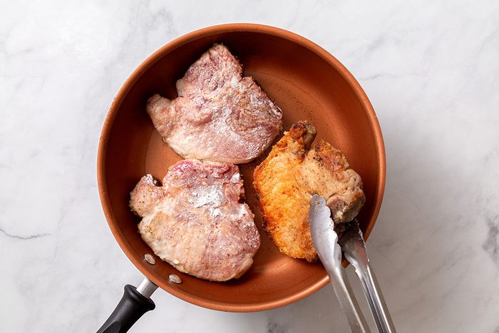 Flour coated pork chops being fried in a large skillet until browned for step two of Smothered Pork Chops in Oven recipe for Taste of Home