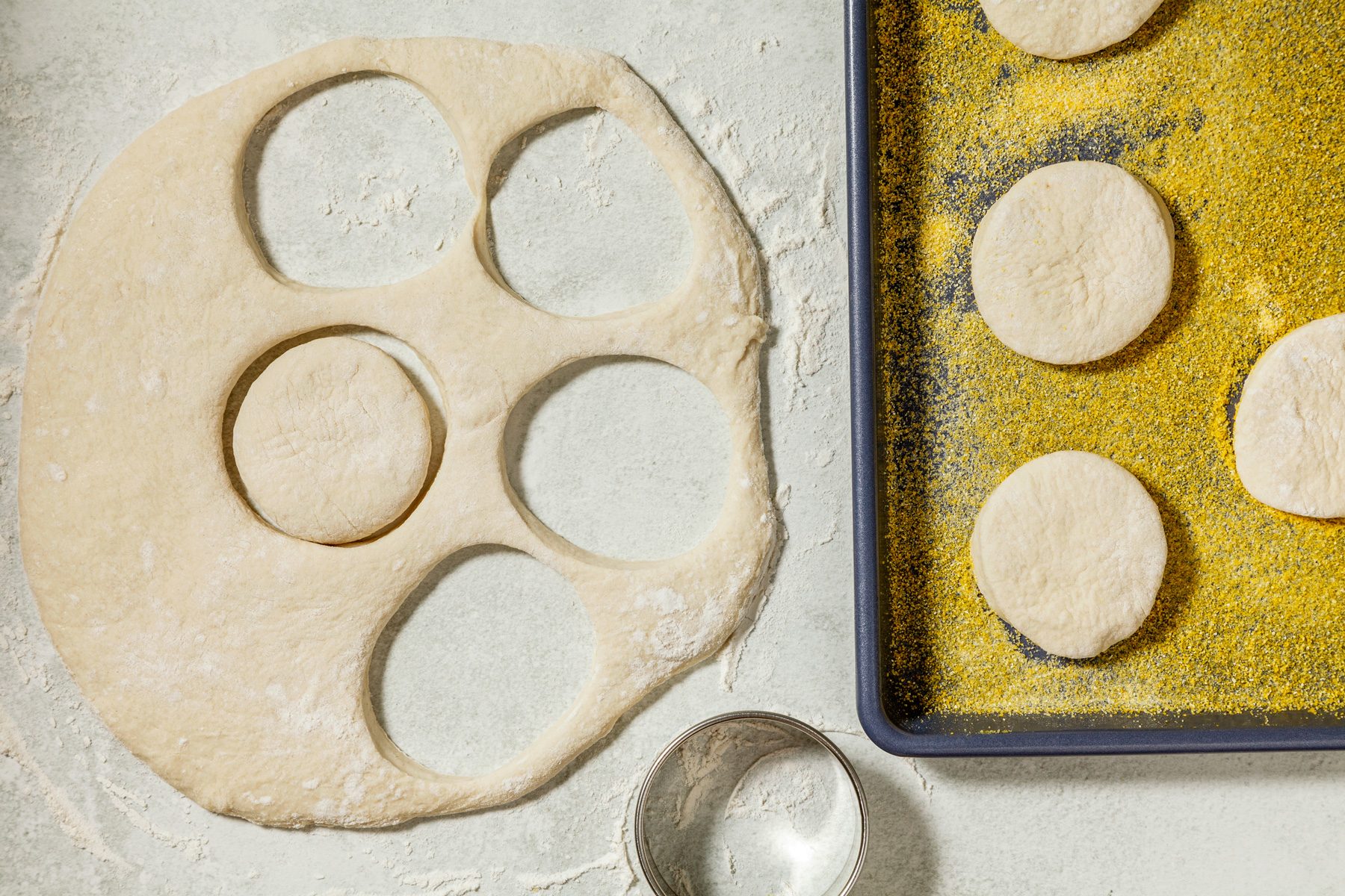 overhead shot of a baking sheet with a few rounds of dough cut from a larger dough circle, The dough is a light yellow color and appears soft and pliable, The dough circles are placed on a bed of cornmeal, A cookie cutter is visible on the surface,