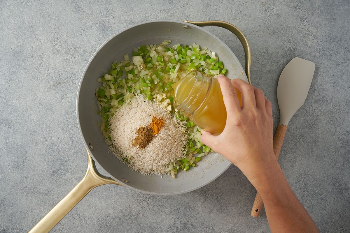 Overhead view of uncooked rice, cumin, turmeric, and chicken broth being added to the skillet along with sauteed veggies for the Taste of Home Southwest Rice recipe.