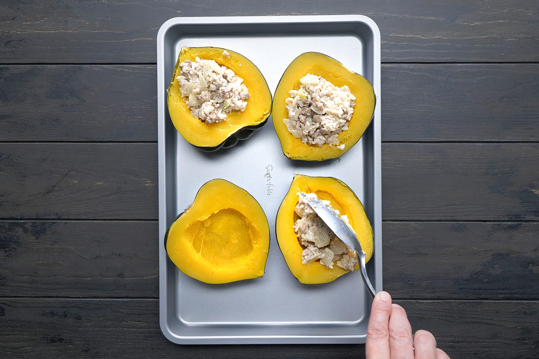 overhead shot of Four acorn squash halves are arranged on a baking sheet, The squash shells are hollowed out and filled with a creamy mixture that appears to contain ground meat, vegetables, and cheese, A spatula is being used to fill one of the squash halves with the mixture;