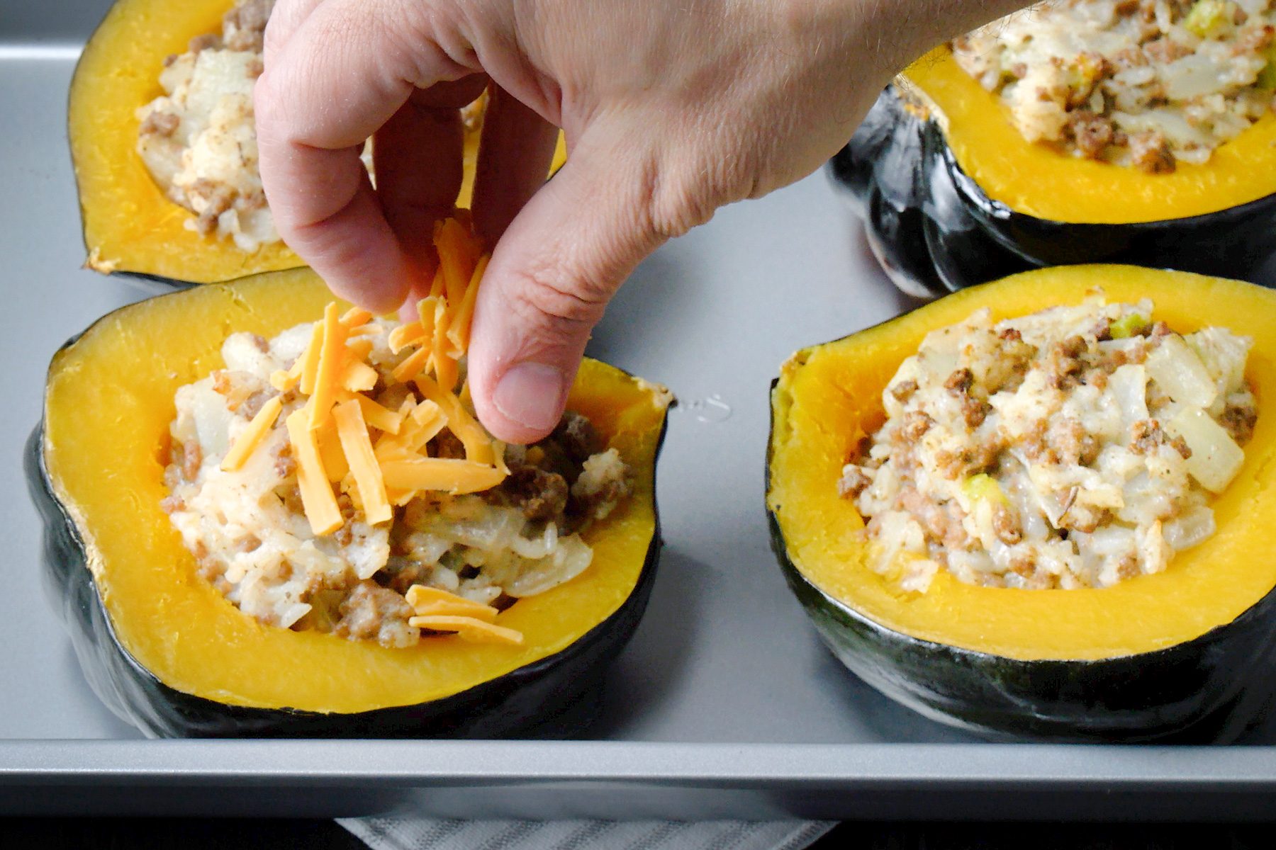 3/4th shot of a baking sheet filled with stuffed acorn squash, A person's hand is sprinkling shredded cheese over one of the stuffed squash halves, The squash shells are a vibrant orange color, and the filling is a mixture of ground meat, rice, and vegetables, The melted cheese is creating a golden brown topping on the squash;