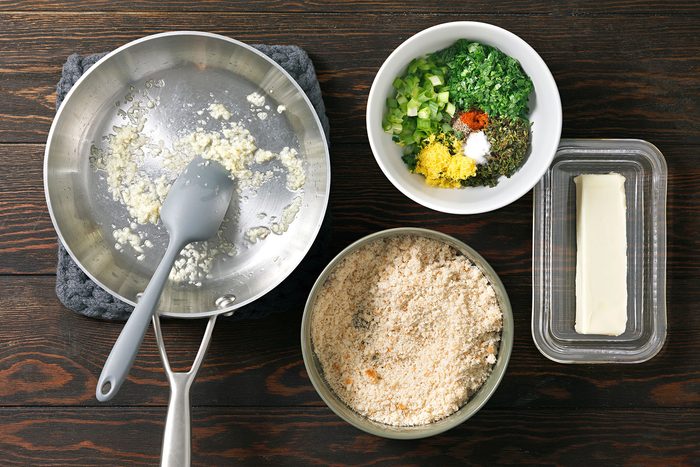 Ingredients for cooking: a pan with sautéed garlic, a bowl of breadcrumbs, a bowl with herbs and spices, and a stick of butter on the side. The items are set on a dark wooden table.