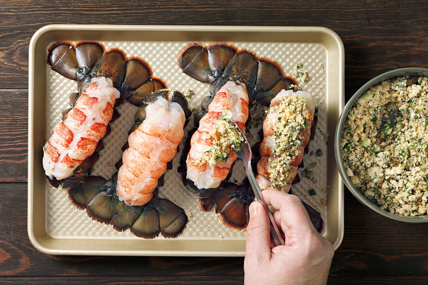 Hand preparing four lobster tails on a baking tray, applying a breadcrumb and herb mixture.