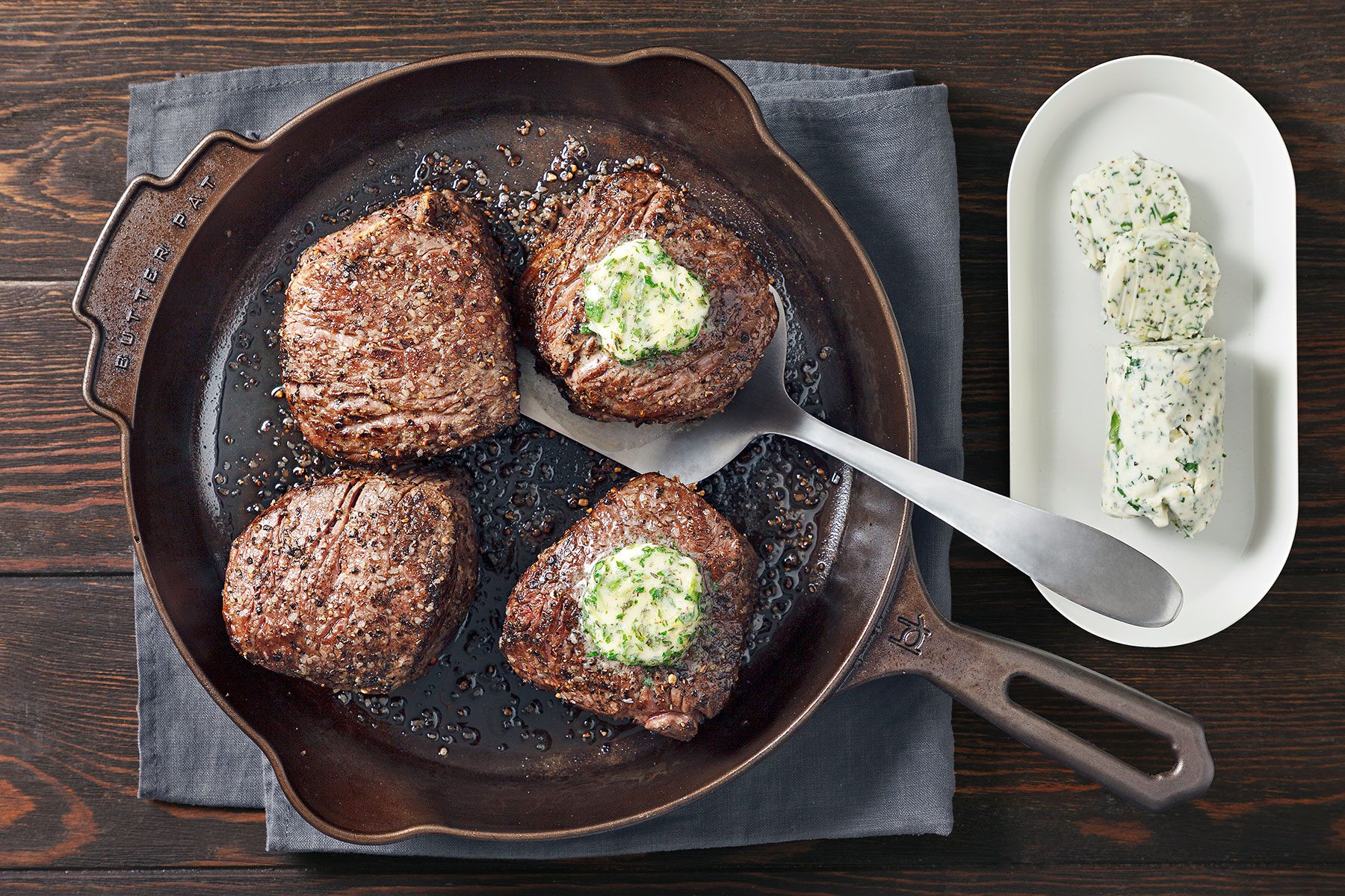 Four grilled steaks in a skillet topped with herb butter, placed on a dark wooden table. A serving knife rests in the skillet. A white plate with additional herb butter slices is beside it.