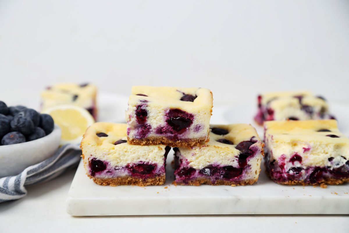 Close up of Taste of Home's Blueberry Cheesecake Bars sliced and served on a platter on a white wooden surface.